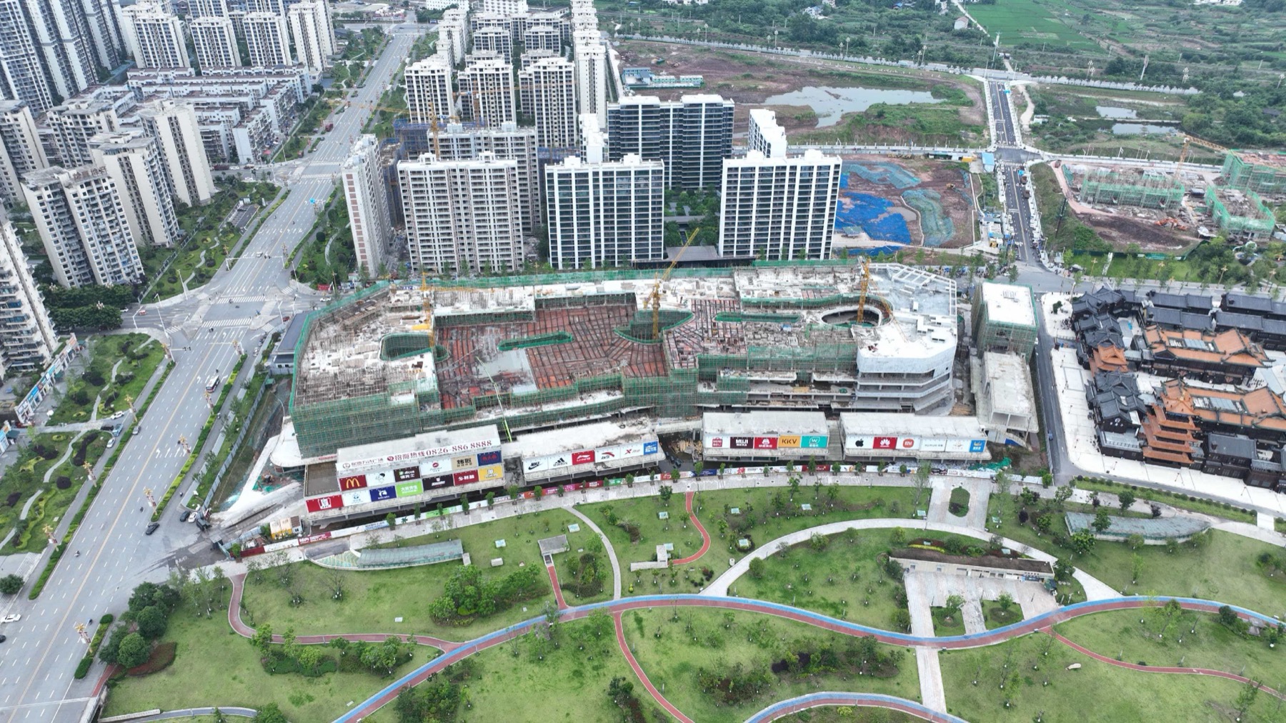 岳池万达广场工地航拍，前景为龙湖公园，画面中可见万达盒子、金街沿街面、塔吊和周边道路